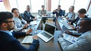 Group of professionals sitting around a conference table during a business meeting, discussing charts and strategies with laptops and documents.