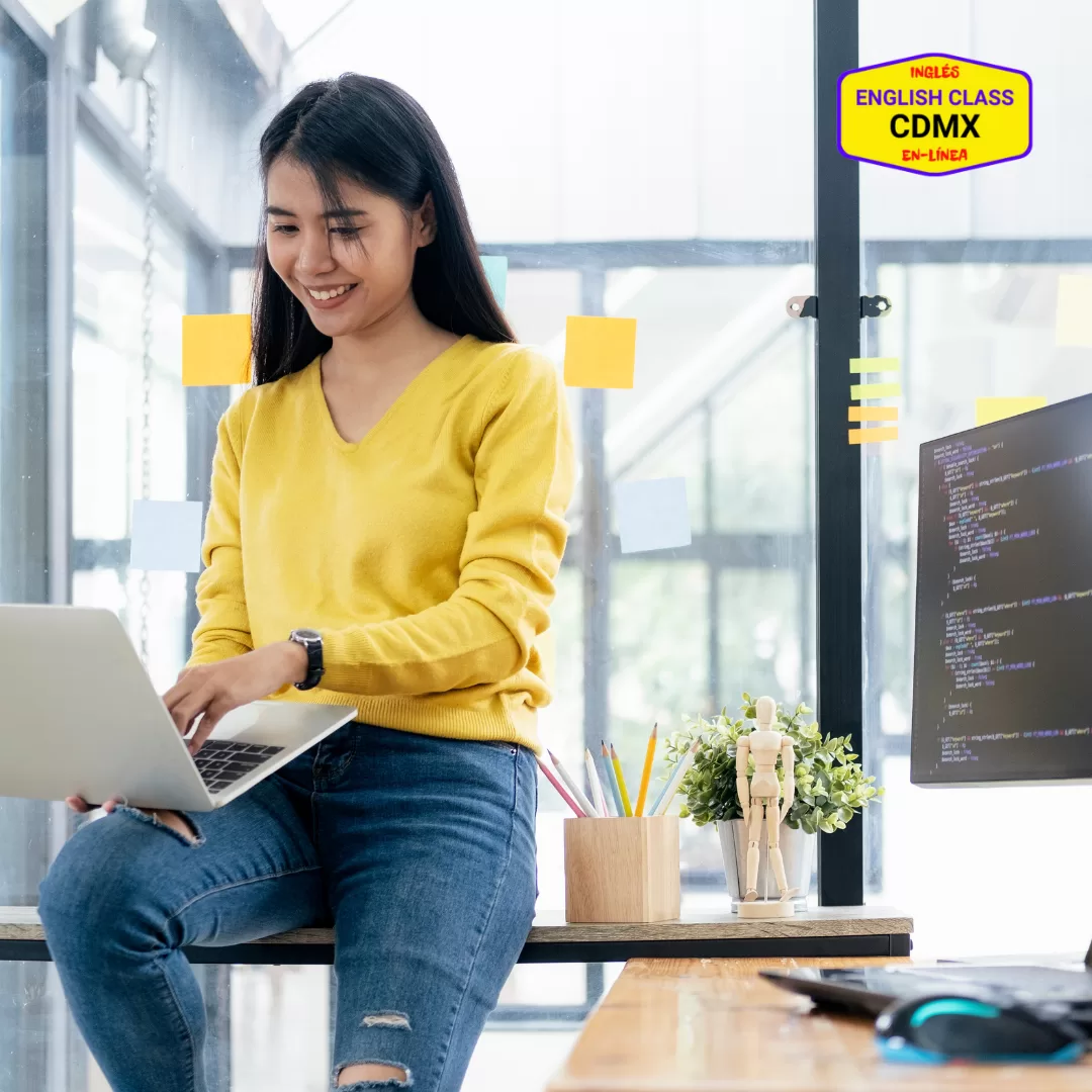 Smiling young woman in a bright workspace using a laptop, with coding on a monitor and the English Class CDMX logo in the background