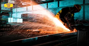 A worker wearing safety gear using a grinding tool in an industrial workshop, with bright sparks flying in the dark environment, featuring the English Class CDMX logo.